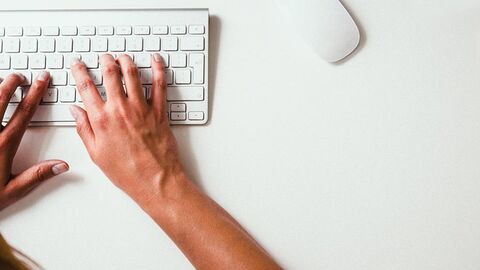 Zwei Hände bearbeiten eine Tastatur, im Hintergrund ein weißer Schreibtisch mit einer Maus.