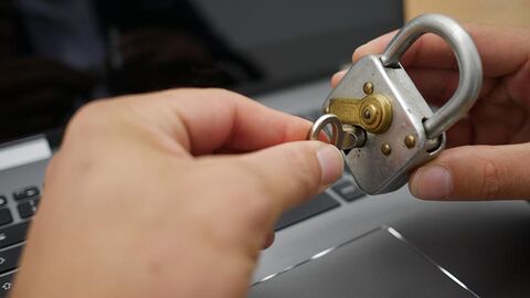 Eine Hand hält ein Vorhängeschloss, die andere schließt es mit einem Schlüssel zu. Im Hintergrund ist eine Tastatur zu sehen.